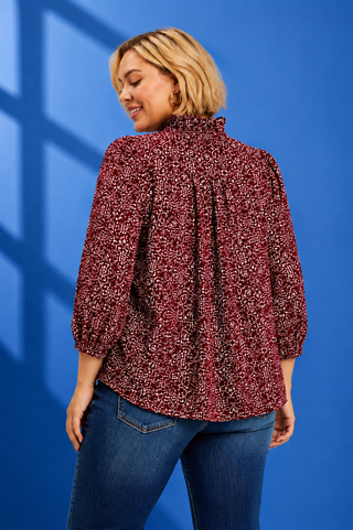 A woman with short blonde hair is shown from the back wearing a dark red and black patterned blouse with three-quarter puff sleeves, paired with blue jeans, against a vibrant blue background with window shadow patterns.