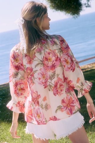 Woman wearing a floral blouse and white shorts standing by a body of water.