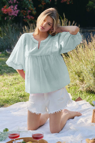 Woman in a light green blouse and white shorts sitting on a picnic blanket in a garden.
