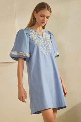 Woman wearing a blue dress with white embroidery against a neutral background