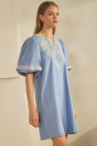 Woman wearing a light blue dress with white embroidery against a beige background