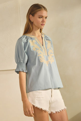 Woman wearing a light blue blouse with yellow embroidery and white shorts against a beige background
