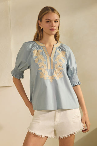 Woman wearing a light blue blouse with gold embroidery and white shorts against a beige background