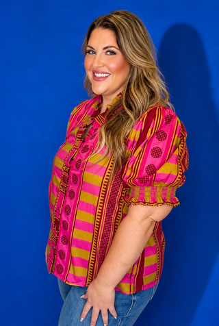 A woman wearing a short-sleeve button-up blouse with a vibrant pink, magenta, and yellow patterned design, paired with blue jeans