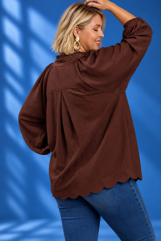 A woman wearing a brown long-sleeve blouse with a scalloped hem and gathered neckline, paired with blue jeans, posing against a blue background with soft shadow overlays.