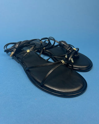 A pair of black leather flat sandals with thin strappy details and small gold hardware, against a solid blue background.