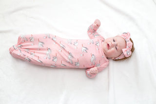 A baby lying on a white blanket wearing a pink footed sleeper with a white and gray ballet dancer pattern and a matching pink bow headband.