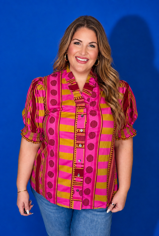 A woman modeling a short-sleeve button-up blouse with a vibrant pink, yellow, and red patterned design, paired with blue jeans