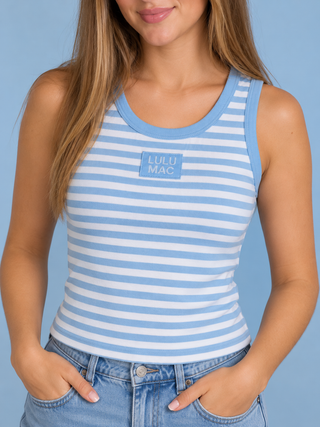 A woman wearing a light blue and white striped tank top with a 'LULU MAC' logo patch on the chest, paired with light-wash blue jeans, set against a solid light blue background.