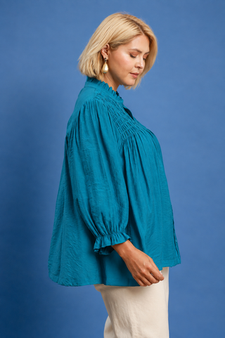 A woman with blonde hair is shown from the side wearing a teal blue long-sleeve blouse with gathered detailing and white pants, standing against a solid blue studio background.