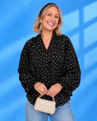 A woman with blonde hair wearing a black long-sleeve blouse with white polka dots, blue jeans, and holding a small beige quilted handbag