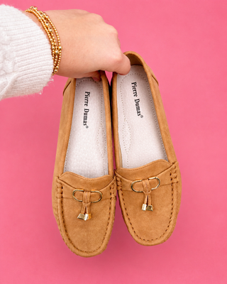 Tan loafers with a gold bow held by a hand against a pink background