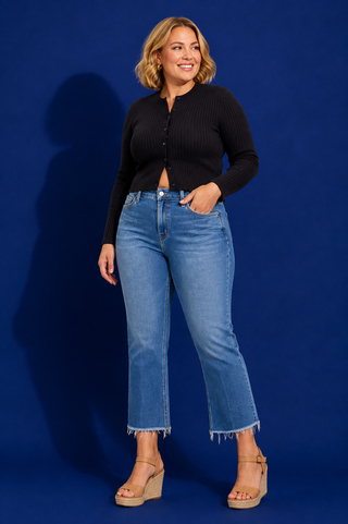 A woman modeling a black ribbed cropped cardigan and blue cropped jeans with frayed hems, paired with beige wedge sandals, set against a solid blue studio backdrop.