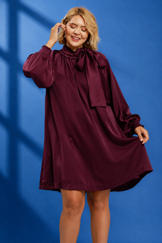 A woman modeling a long-sleeved, high-neck satin dress in deep burgundy with a large bow at the collar, posed against a blue background with soft window shadow overlays.