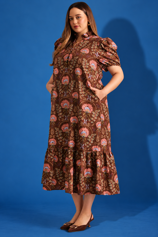A woman modeling a brown bohemian-style midi dress with puff sleeves, a high ruffled collar, and an all-over floral print in pink, orange, and blue