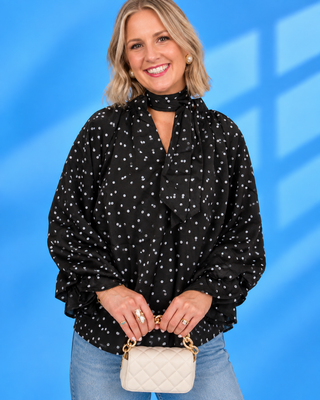 A smiling blonde woman wearing a black long-sleeve blouse with a white geometric pattern, a high neck choker detail, and ruffled trim