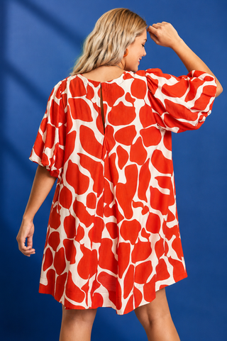 A woman shown from the back wearing a loose, short-sleeve dress with a bold red and off-white abstract print, posed against a solid blue studio background.