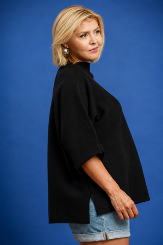 A blonde woman wearing a black oversized mock-neck top and light blue denim shorts, posing against a solid blue studio background.