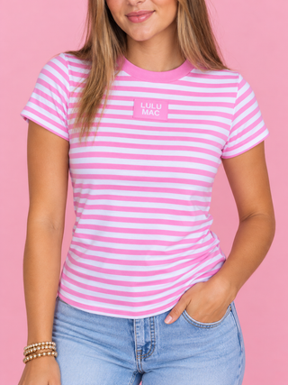 A woman wearing a pink and white striped short-sleeve t-shirt with a pink 'LULU MAC' logo on the chest, paired with light blue jeans, set against a solid pink background.