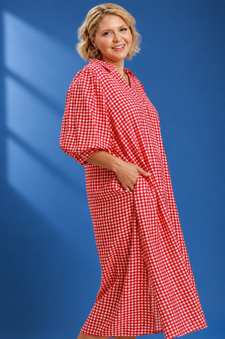 A woman modeling a red and white gingham long dress with three-quarter sleeves and side pockets, posed against a solid blue studio background.