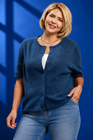 A woman with blonde hair wearing a short-sleeve blue knit cardigan over a white top and blue jeans, standing against a blue background with windowpane shadows.