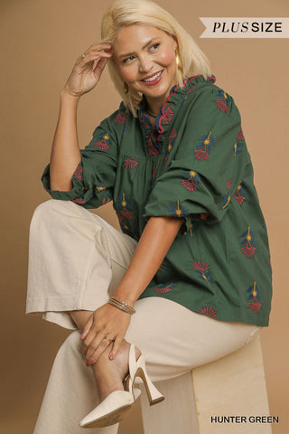 Woman wearing a green blouse with floral patterns and beige pants, sitting against a brown background.