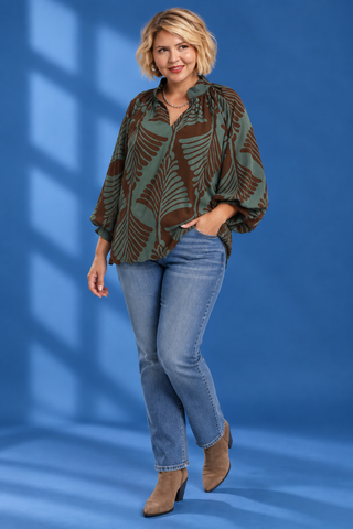 A woman modeling a green and brown tropical leaf print blouse with blue jeans and brown ankle boots, standing against a blue studio backdrop with window shadow overlays.