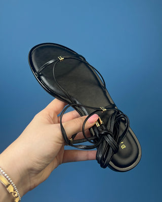A pair of black leather flat sandals with thin strappy details and small gold hardware, against a solid blue background.
