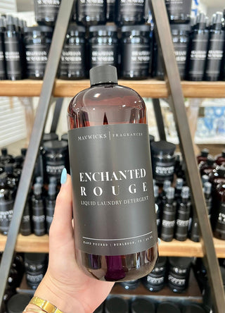 Hand holding a bottle of Maywicks Enchanted Rouge laundry detergent with shelves in the background.