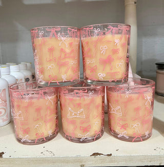 Four transparent candles with pink bow designs in clear containers on a shelf.