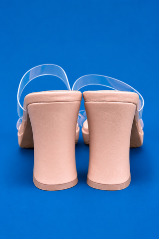 A pair of nude beige block-heel mule sandals with transparent straps, shown from the back against a solid blue background.