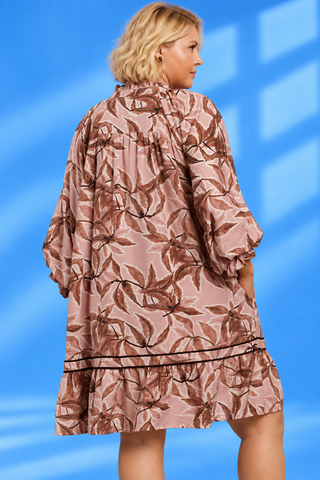 A woman wearing a loose, short-sleeve kaftan-style dress with a brown leaf pattern on a dusty pink background
