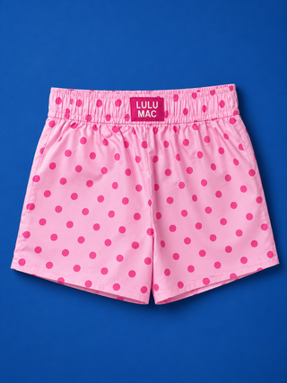 Pink swim trunks with a magenta polka dot pattern and an elastic waistband featuring a pink label that reads 'LULU MAC'. The shorts are laid flat on a solid blue background.