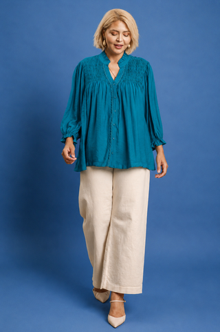 A woman modeling a teal long-sleeve blouse with a gathered, smocked-style neckline and buttons down the front, paired with wide-leg cream trousers, against a solid blue studio background.