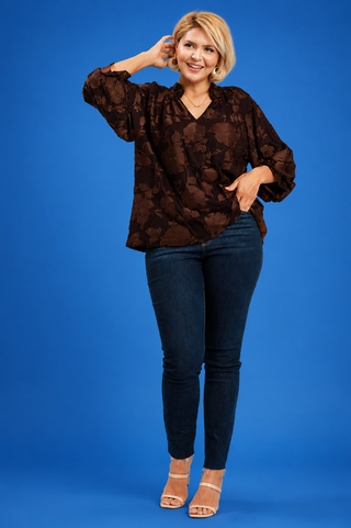 Woman wearing a brown patterned blouse and dark jeans against a blue background