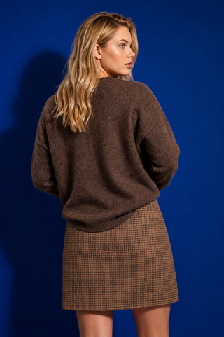 A woman with blonde hair is shown from the back wearing a brown fuzzy knit sweater and a brown and beige houndstooth mini skirt, set against a solid blue studio background.