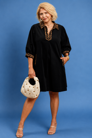 A woman modeling a black short-sleeve tunic dress with gold embroidered trim on the collar, cuffs, and neckline, holding a cream-colored studded handbag against a solid blue studio background.