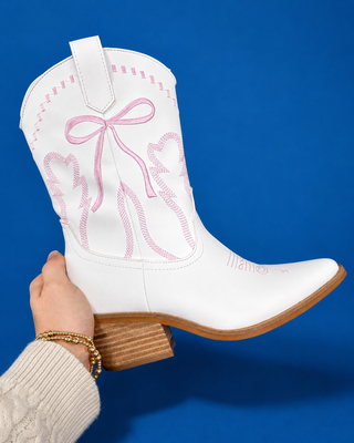 A pair of white leather cowboy boots with pink embroidered bow and leaf designs, displayed on a solid blue background.