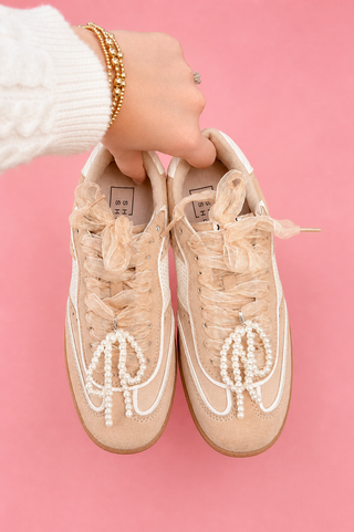 Beige sneakers with pearl details held by a person against a pink background