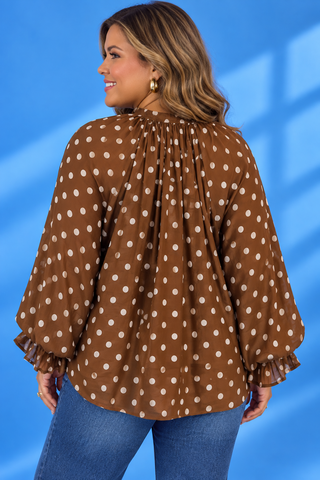 A woman wearing a brown long-sleeve blouse with white polka dots and ruffled cuffs, paired with blue jeans