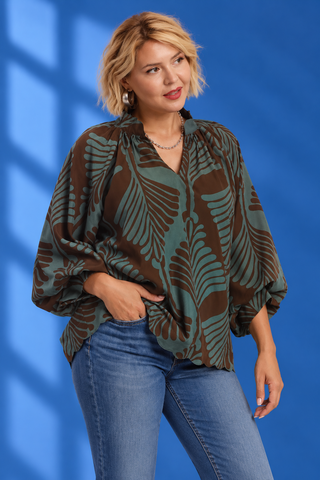 A woman modeling a loose, long-sleeve blouse with a brown and sage green palm leaf pattern, paired with blue jeans