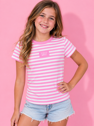 A young girl with long light brown hair smiling, wearing a pink and white striped t-shirt with a small pink logo on the chest, paired with light blue denim shorts, set against a solid pink background.