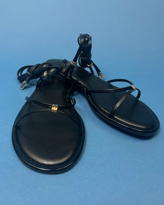 A pair of black leather flat sandals with thin strappy details and small gold hardware, against a solid blue background.