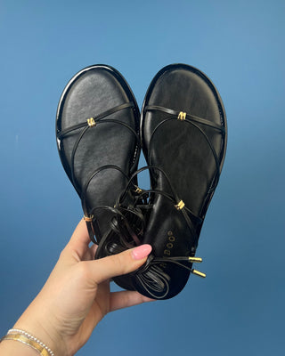 A pair of black leather flat sandals with thin strappy details and small gold hardware, against a solid blue background.