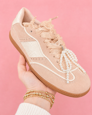 Beige sneaker with pearl laces held by a hand against a pink background