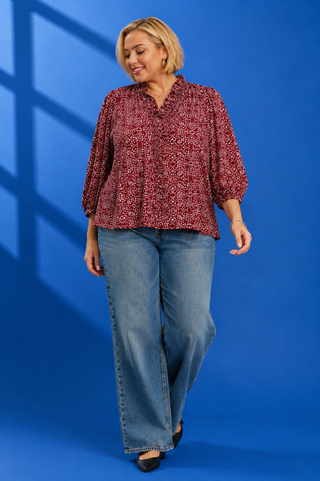A woman modeling a red and white patterned blouse with voluminous sleeves, paired with blue jeans, against a solid blue studio backdrop with window shadow overlays.