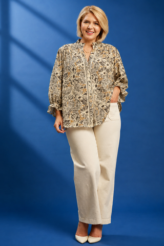 A woman modeling a long-sleeve button-up blouse with an abstract botanical pattern in beige, brown, and black, paired with cream trousers and white heels, set against a blue studio backdrop.