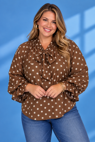 A woman wearing a brown long-sleeve blouse with white polka dots and a tie-neck bow, paired with blue jeans