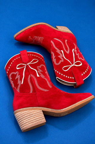 A pair of red suede cowboy boots with white embroidered western patterns and bows, displayed on a solid blue background.