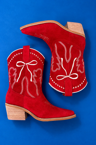 A pair of red suede cowboy boots with white embroidered western-style patterns and bows, displayed on a solid blue backgroun